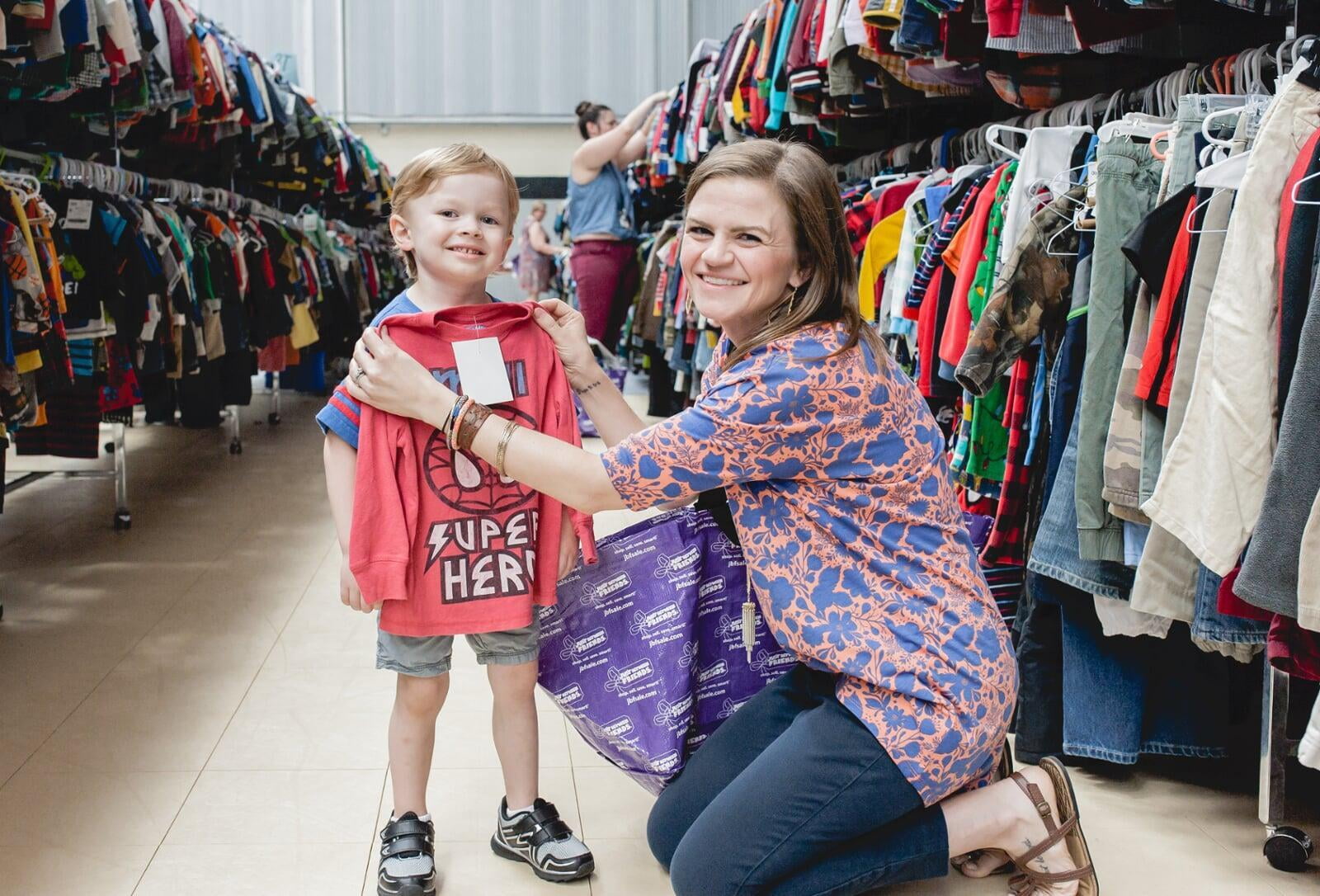 Mom wears baby and holds toddler boy clothes in each hand. Second mom shops behind her from the racks of boys clothes.